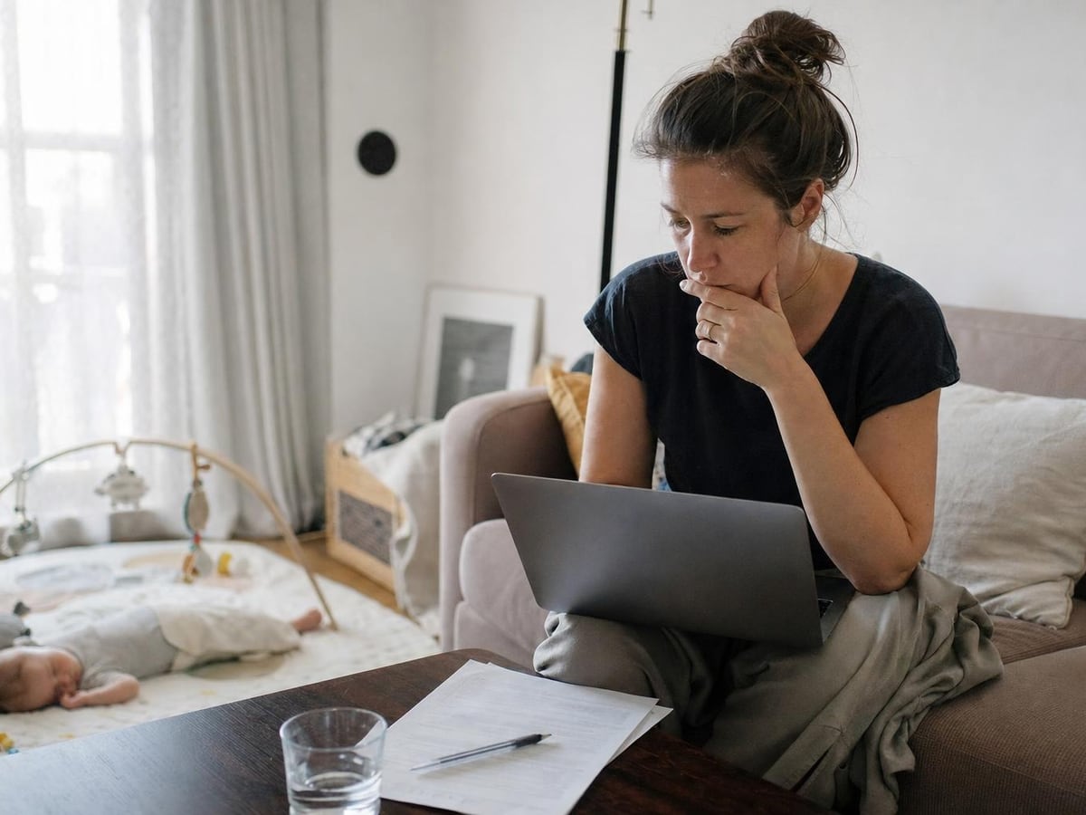 a mother on parental leave weighs a daycare form at her laptop, her baby asleep nearby