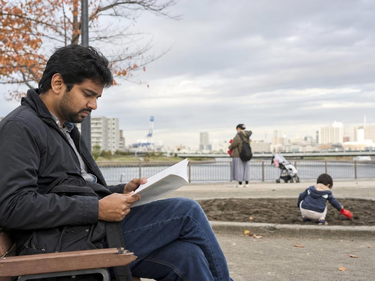 a father reads a daycare guide on a park bench while his toddler plays in the sand